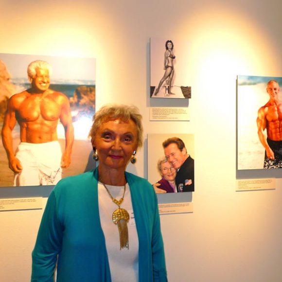 Bodybuilder Doris Barrilleaux at the 2011 Opening of the Joe and Betty Weider Museum of Physical Culture; a photograph of Barrilleaux posing and another of her with bodybuilder Arnold Schwarzenegger are two of the four photographs in the background.