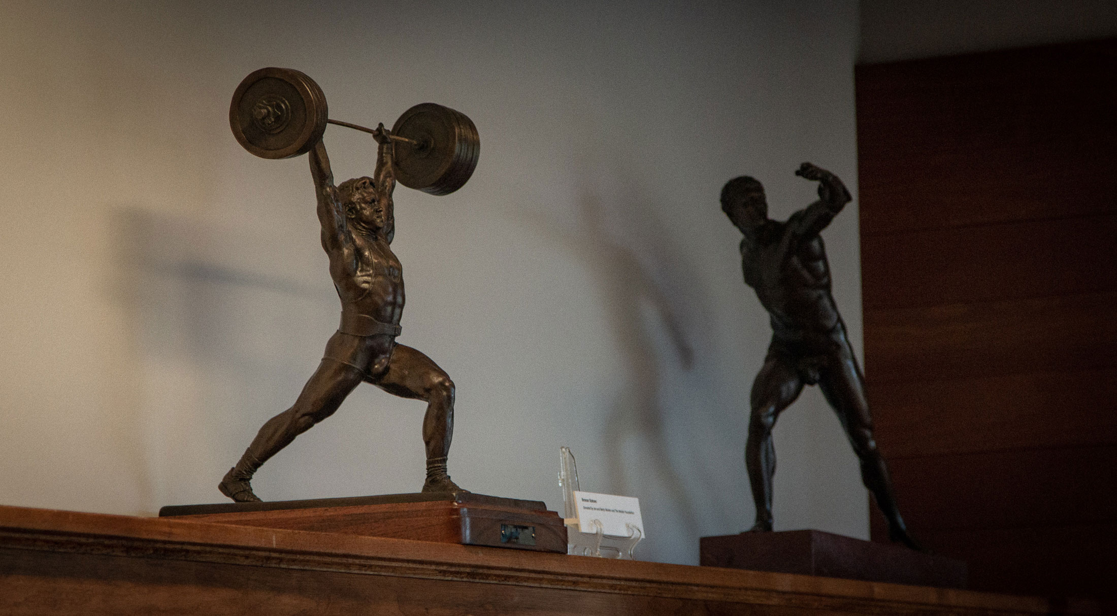 Two physical culture sculptures: a weightlifter pressing a barbell overhead with two hands, and a discus thrower, from the Weider Art Collection, in the Reading Room.
