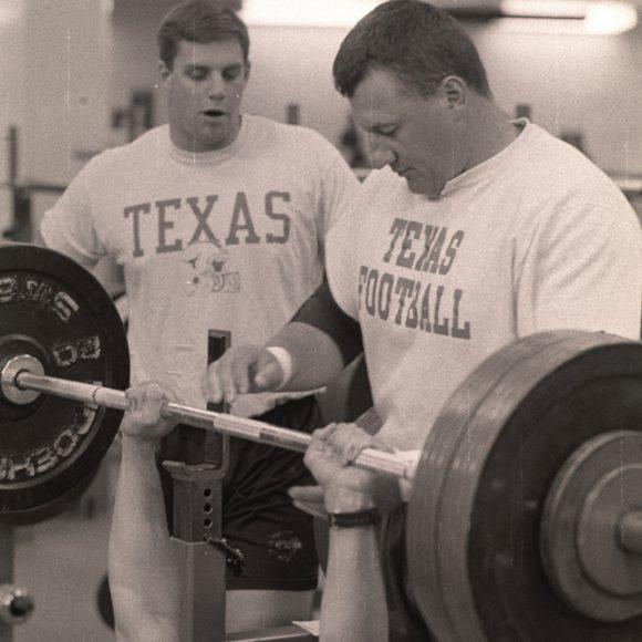 Photograph of University of Texas at Austin strength training showing a Texas football player about to bench press a barbell while two others spot him.
