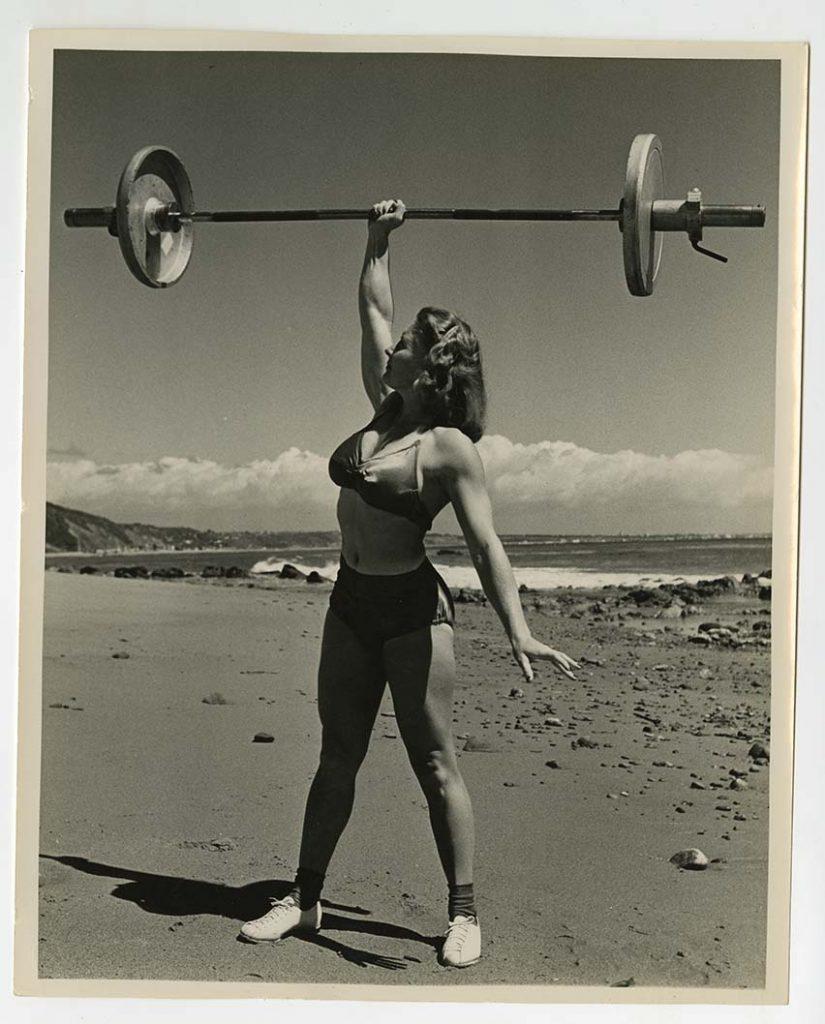 Bodybuilder Pudgy Stockton pressing a barbell overhead with one hand, in a bkini, on the beach, from the Pudgy and Les Stockton Collection.