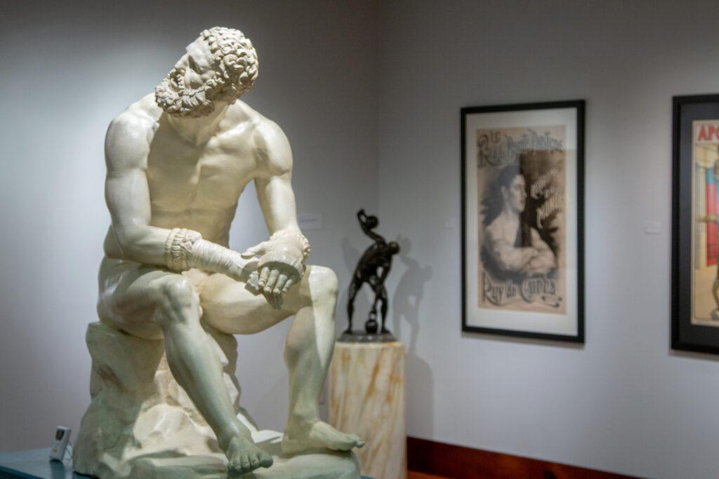 The Battle Cast of the Boxer, on loan from the Blanton Museum of Art, in the Teresa Lozano Long Art Gallery, in the main lobby; a sculpture and a poster of strongman Ruy da Cunha are in the background.