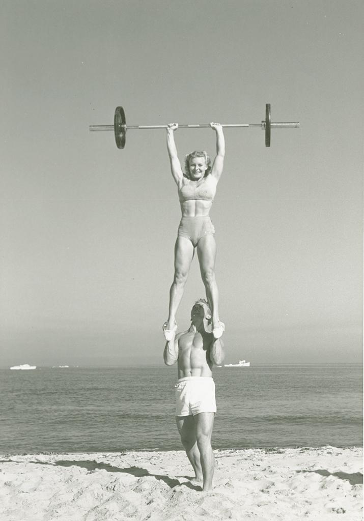 Bodybuilder Les Stockton, in a swimsuit, balancing bodybuilder Pudgy Stockton on his hands while she presses a barbell overhead with two hands, in a bikini, on the beach, from the Pudgy and Les Stockton Collection.