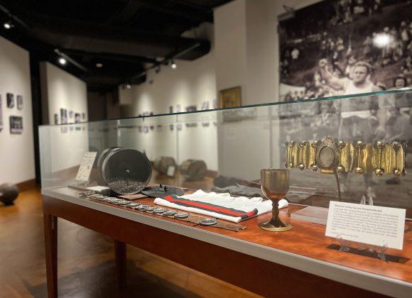A photograph of the glass display case in the Strong Men, Strong Women exhibit. The contents of the case are a milo adjustable barbell, Donald Dinney's belt, Jack Shanks' lifting vest, and Minerva's loving cup and championship belt.