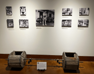 A photograph of a wall within the Strong Men Strong Women exhibit. The focal photo is of Bob Peoples and below that photo is an artifact, Bob People's wooden barbell.
