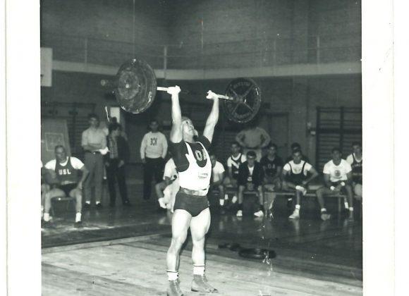 Weightlifter Bob Samuels pressing a barbell overhead with two hands, while nine other weightlifters look on, from the Bob Samuels Collection.