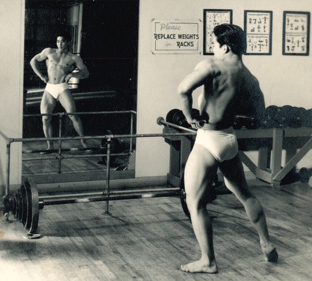 Tommy Kono poses in a mirror as a bodybuilder.