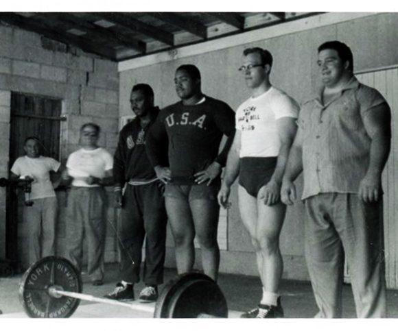 John Davis, Jim Bradford, Norbert Schemansky, and Paul Anderson of the US Weightlifting Team stand behind a barbell.