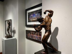 A bronze sculpture of Arnold Schwarzenegger posing in front of two photos and another sculpture in the new Arnold Sport Festival exhibit.