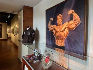 This photograph shows the entrance to the Arnold & Jim exhibit. An oil painting of Arnold Schwarzenegger posing is the central focus but artifacts in a glass case and a jacket are also visible.