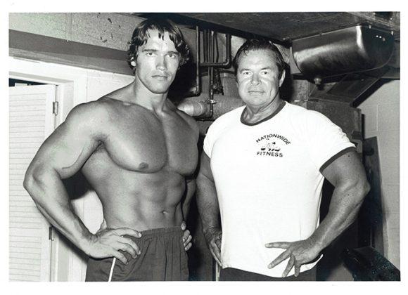 A black and white photograph of Arnold Schwarzenegger (shirtless) standing with friend and business partner Jim Lorimer.