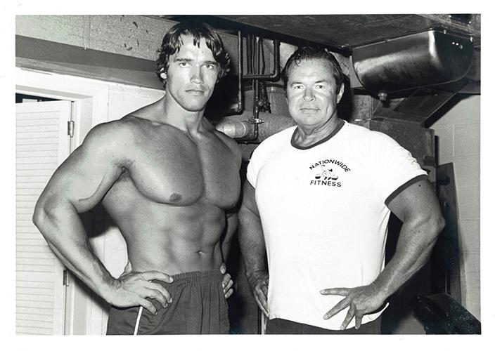 A black and white photograph of Arnold Schwarzenegger (shirtless) standing with friend and business partner Jim Lorimer.