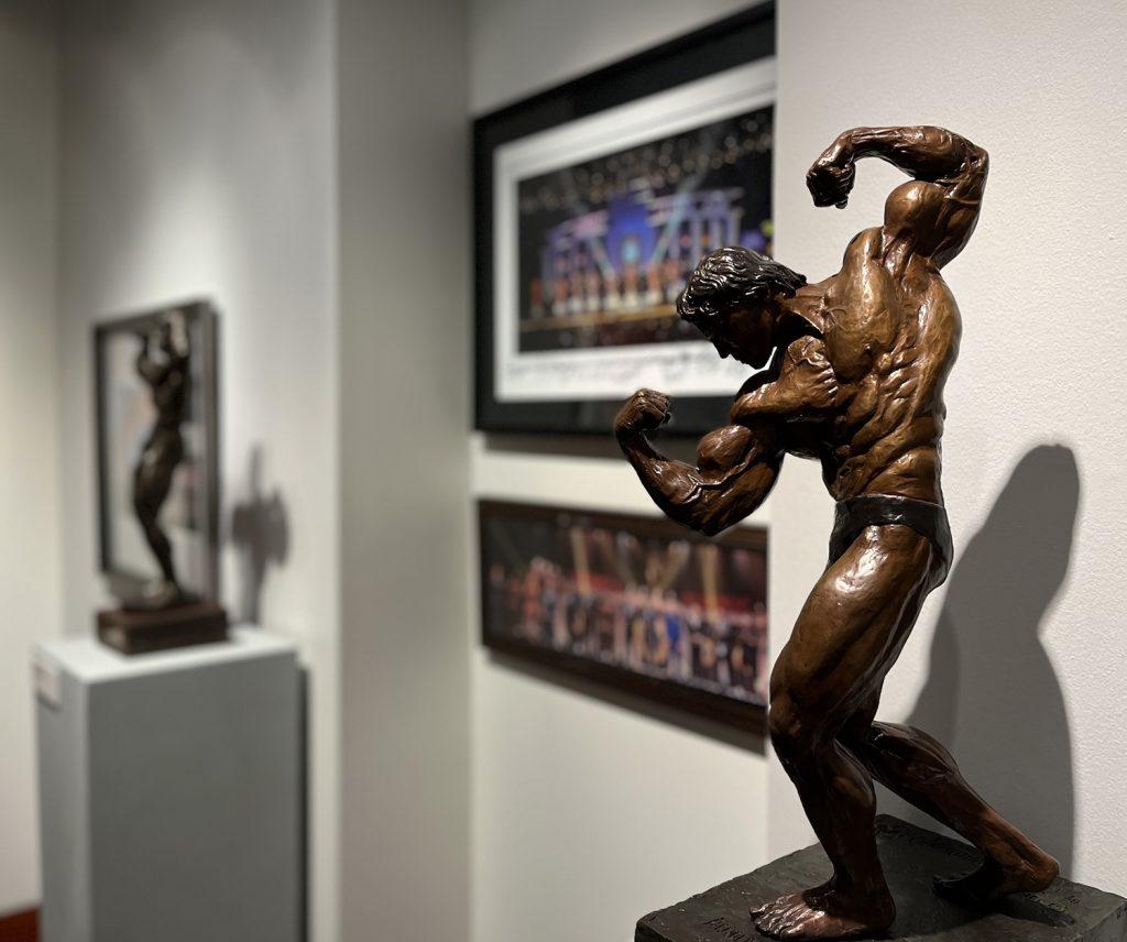 A bronze sculpture of Arnold Schwarzenegger posing in front of two photos and another sculpture in the new Arnold Sport Festival exhibit.