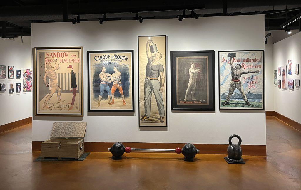 Five lithographic posters with 3 weight lifting artifacts displayed on the floor below them.