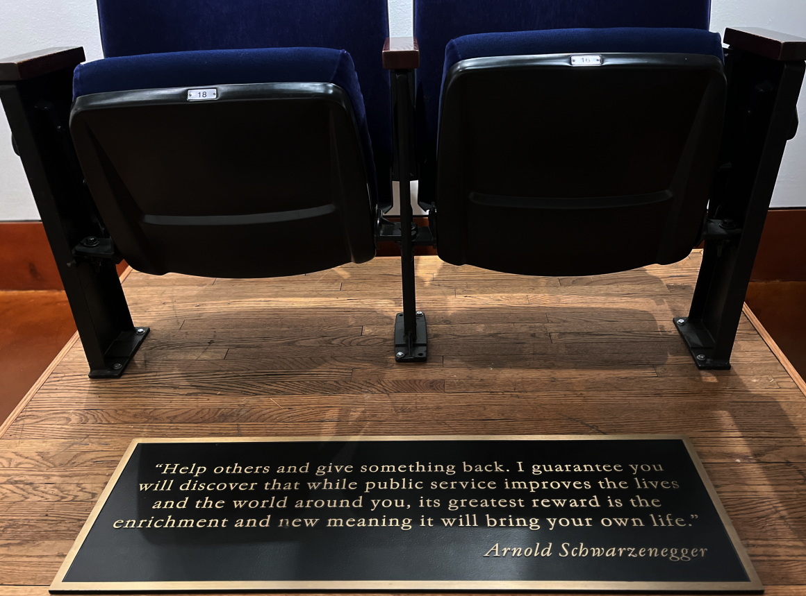 Two classic theatre seats with blue upholstery. A black plaque with bronze lettering features a quote by Arnold Schwarzenegger, reads "Help others and give something back. I guarantee you will discover that while public service improves the lives and the world around you, its greatest reward is the enrichment and new meaning it will bring your own life."