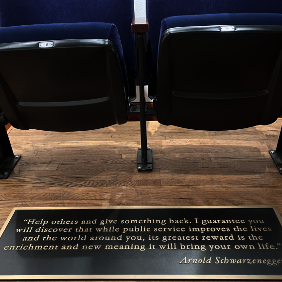 Two classic theatre seats with blue upholstery. A black plaque with bronze lettering features a quote by Arnold Schwarzenegger, reads "Help others and give something back. I guarantee you will discover that while public service improves the lives and the world around you, its greatest reward is the enrichment and new meaning it will bring your own life."
