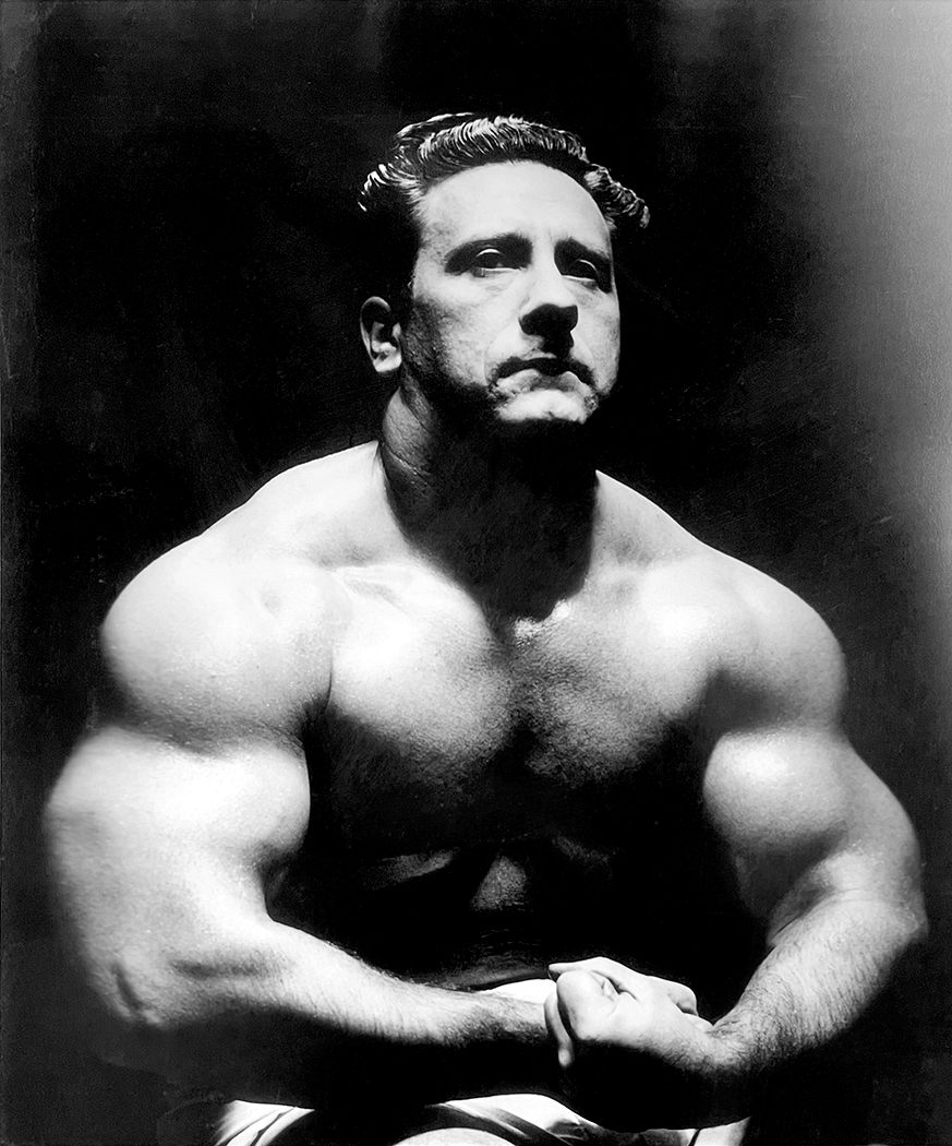 A photo of Joe Weider as a young man. He is over two hundred pounds and very muscular, flexing both his arms.