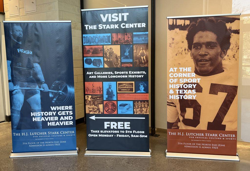 Three new retractable banners on display in the NEZ lobby. From left to right: first is blue tinted with Hafthor Bjornsson deadlifting, second is a blue red and yellow tinted images of Stark highlights and the text "Visit The Stark Center," and third is a burnt orange tinted banner featuring Julius Whittier in his #67 football jersey.