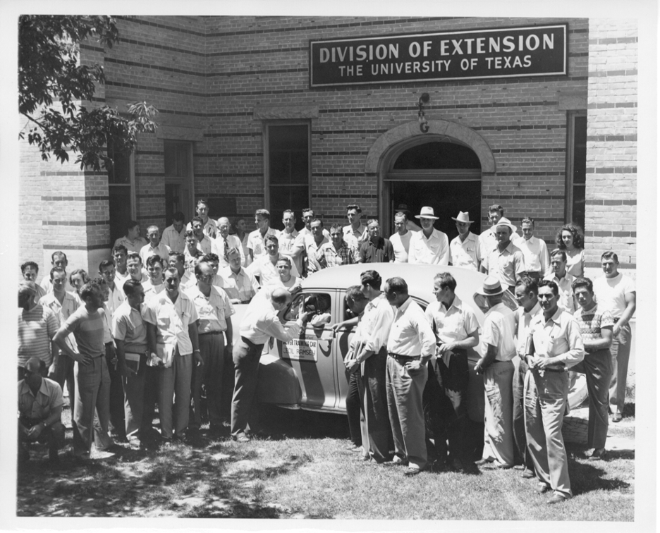 A drivers education course meets outside to gather around a VW Beetle in 1956 on the University of Texas campus.