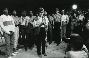 A black and white photograph of University of Texas Longhorn women's basketball coach, Jody Conradt. Conradt stands center holding a microphone with her players in a row behind her, many photographers and camera operators record what she is saying.