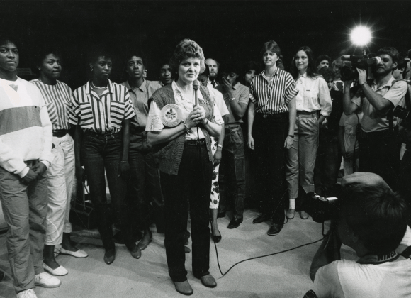 A black and white photograph of University of Texas Longhorn women's basketball coach, Jody Conradt. Conradt stands center holding a microphone with her players in a row behind her, many photographers and camera operators record what she is saying.