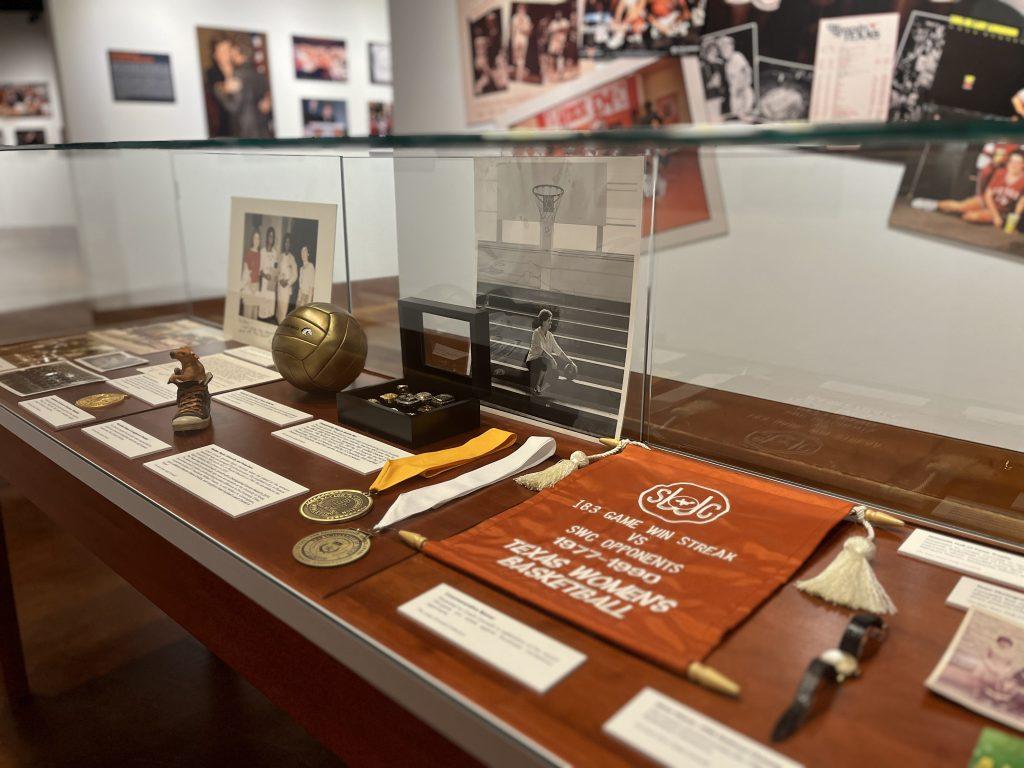 A photo of a table style display case in which several small artifacts from Jody Conradt's personal collection are on display. A burnt orange banner, two medals, photo prints, a jewelry box, a small volleyball, and more.