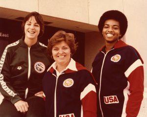 A photo of three women in track suits or warmups outside the Frank Erwin Center. In center is Coach Jody Conradt. This is for Team USA.