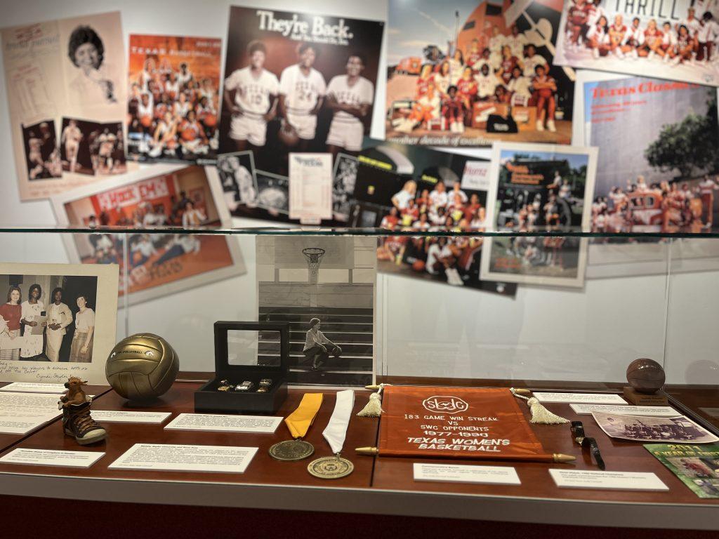 A photo of a table style display case in which several small artifacts from Jody Conradt's personal collection are on display. A burnt orange banner, two medals, photo prints, a jewelry box, a small volleyball, and more.