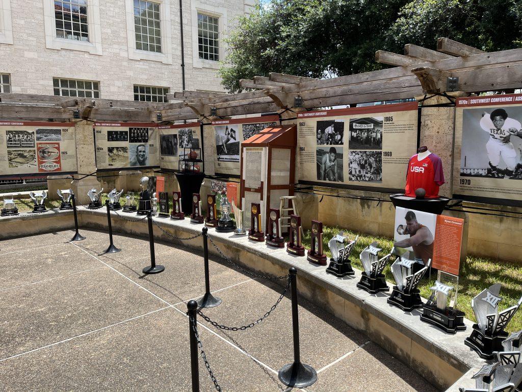 A display of all trophies won by University of Texas varsity sports in the 2023-2024 seasons sit on a curved cement bench. Behind those trophies are a display of artifacts: 3 commemorative basketballs from Jody Conradt's collection, Ricky Williams' wood football locker, and Ryan Crouser's Team USA shot put jersey.