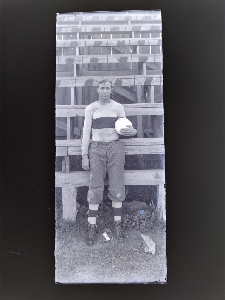 1910 photo of Arnie Kirkpatrick in his uniform holding a football in his elbow in front of the stands.