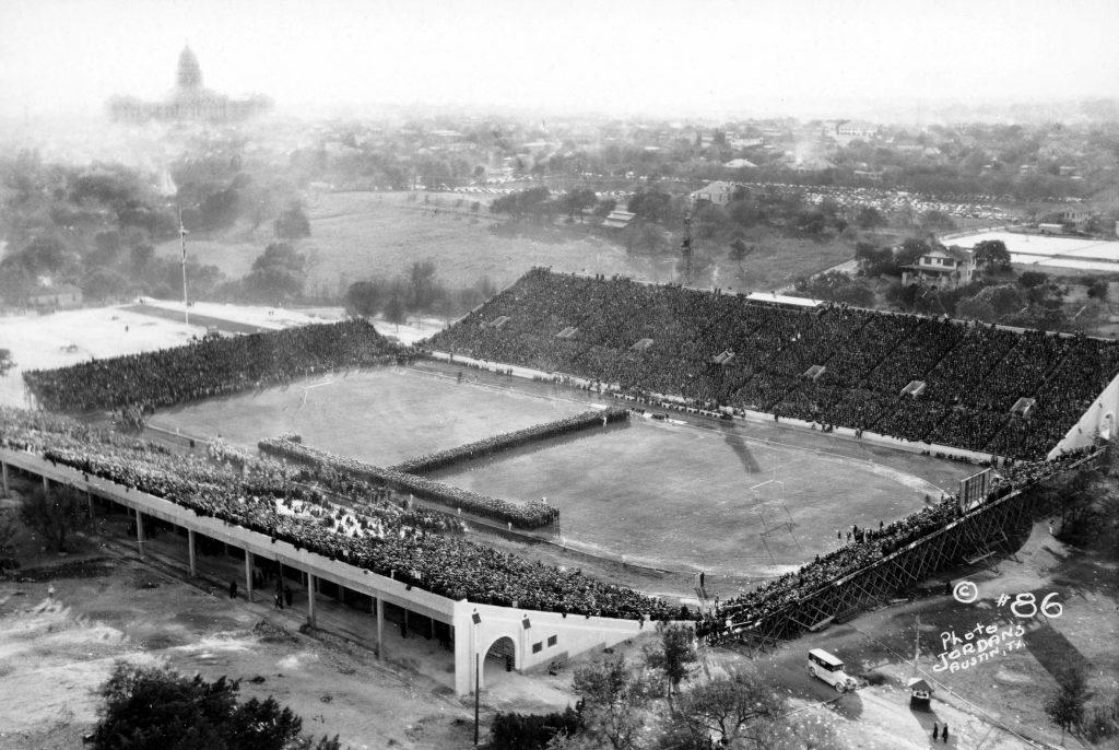 A black and white aerial photo of Texas Memorial Stadium in 1924 during the dedication game. A band or formation of people in the iconic Texas 'T' at center field.