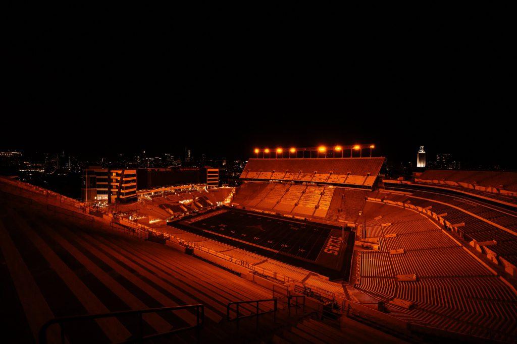 A night time photograph of DKR Texas Memorial Stadium while the burnt orange LEDs flood the field and stands in orange light.