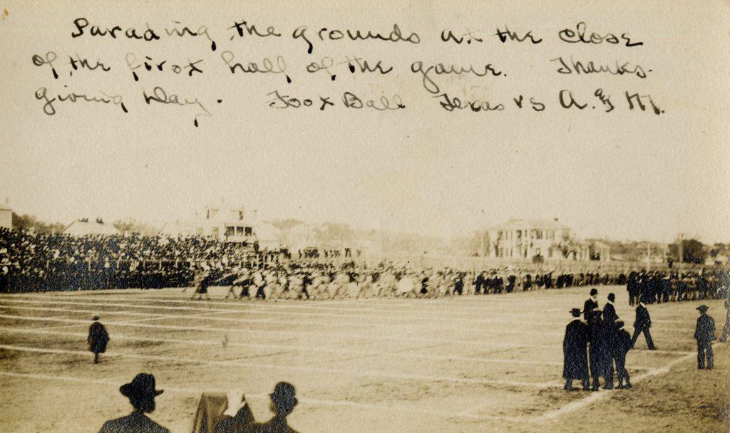 A sepia toned photograph shows a parade of people, maybe a marching band, performing on Clark Field at half time of the 1906 football game between Texas and A&M.