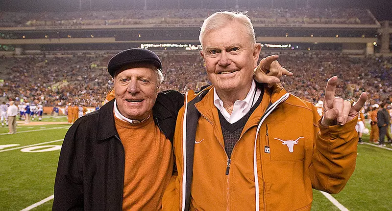 Joe Jamail and Darrell K Royal stand on the sidelines of a University of Texas football game and pose for a photo together by holding up the hook em hand gesture.