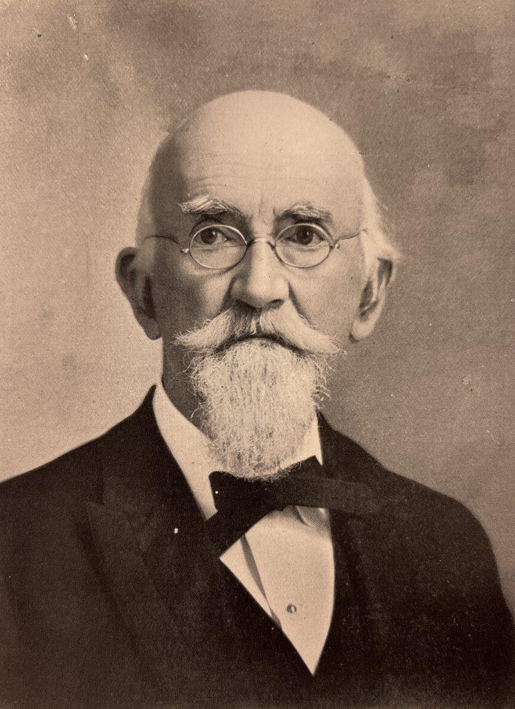 A black and white portrait of an elderly white man who is mostly bald with a long goatee and wearing a bow tie and suit.