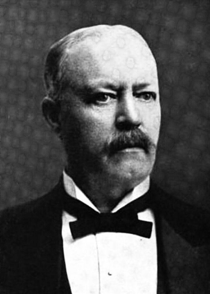 A black and white photo of a middle-aged white man in bow tie and jacket. He is balding and has a mustache.