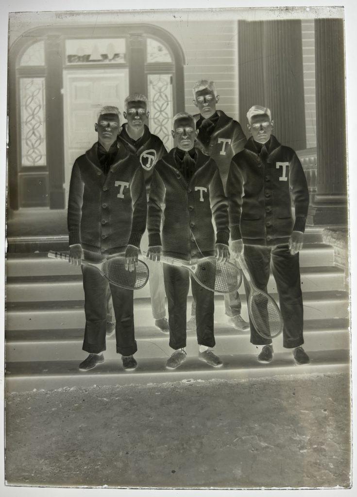 A glass positive plate of the five members of the 1914 University of Texas Men's tennis team holding rackets and wearing their letterman sweaters.