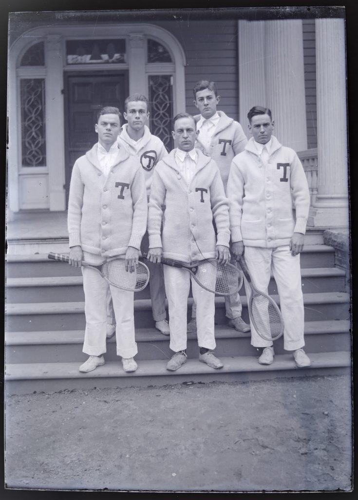 The five members of the 1914 men's tennis team in their off-white letterman sweaters holding tennis rackets in front of a building.