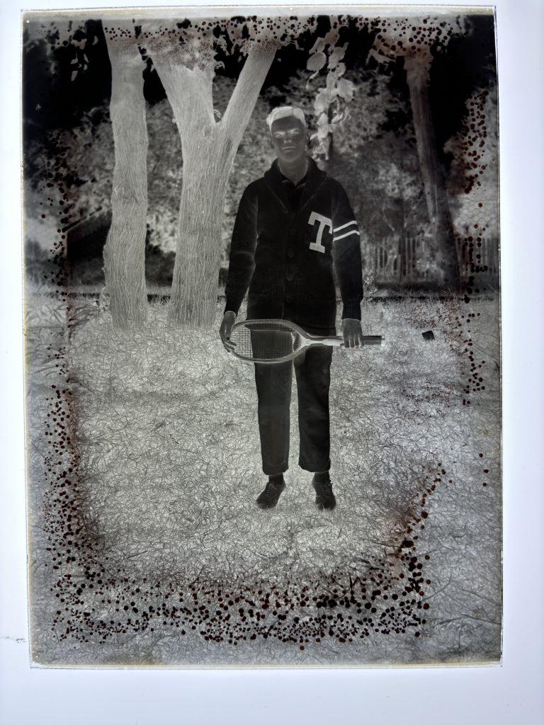 The glass positive plate of 1914 men's tennis player Gillespie Stacy in his letterman's sweater and holding a tennis racket in which the colors are reversed.