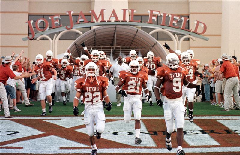 The Texas Longhorn football team, wearing burnt orange jerseys, run out of the South end zone tunnel underneath big letters that read, "Joe Jamail Field."