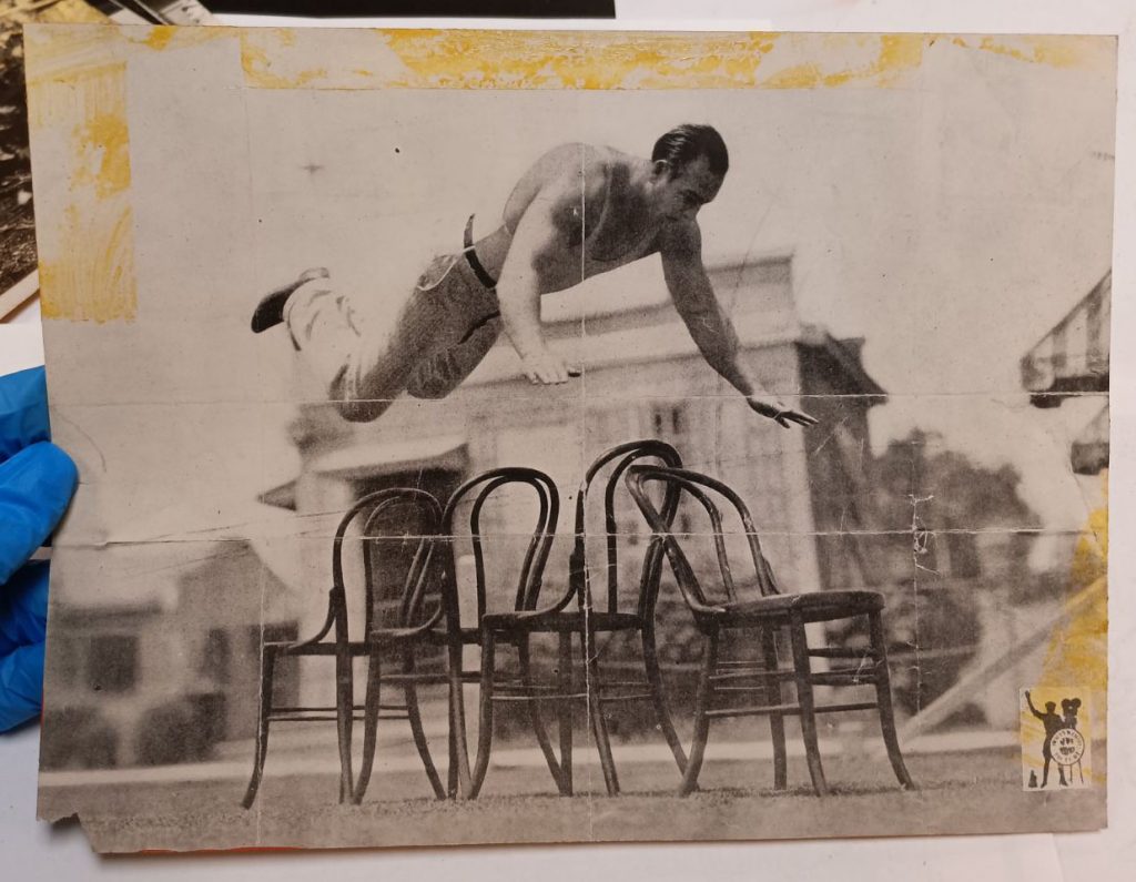 Black and white photograph of Joe Bonomo jumping head first over 4 chairs. The photograph has yellow stains and residue around the top and sides and red paint on the bottom.