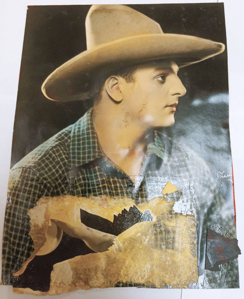 Hand colored photograph of Joe Bonomo in plaid shirt and cowboy hat. The photograph has red paint on the front and sides. The front also has a piece of black construction paper and fragments of another photograph stuck to it.