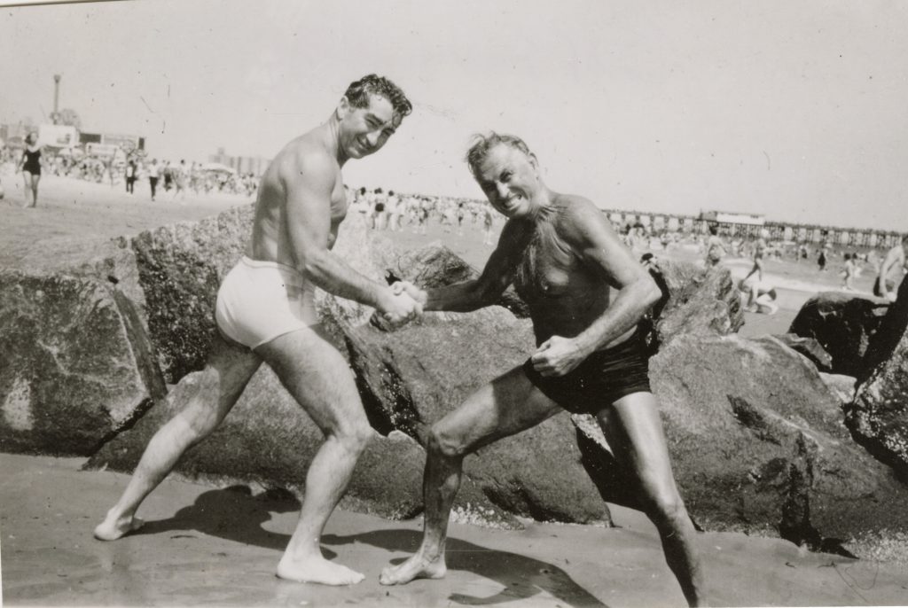 Black and white photograph of Vic Boff and Paul Bragg in swimsuits hand wrestling on Coney Island Beach. They are standing in front of large rocks with beach goers and the pier in the background. 