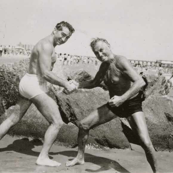 Black and white photograph of Vic Boff and Paul Bragg in swimsuits hand wrestling on Coney Island Beach. They are standing in front of large rocks with beach goers and the pier in the background.