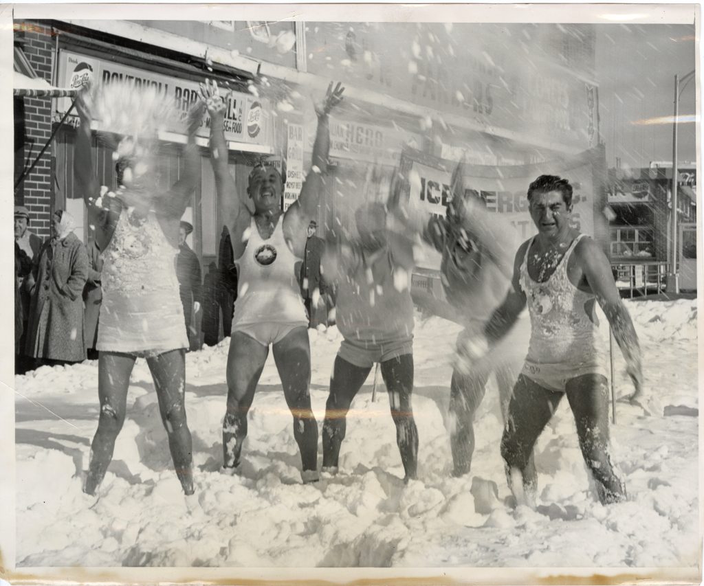 Black and white photograph of Vic Boff and four other members of the Iceberg Athletic Club. All five men are in white swim trunks and tank tops, throwing snow up in the air. Behind them are people in coats and hats, storefronts, and a banner for the Club. 