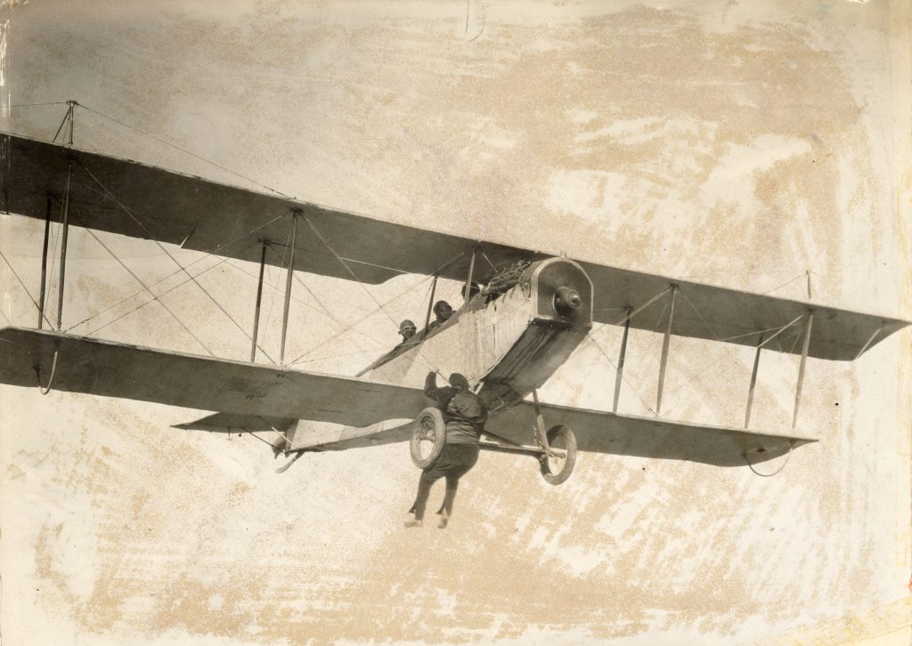 Black and white photograph of Joe Bonomo hanging on to the front tires of an airplane in mid flight. Three men are in the plane. 