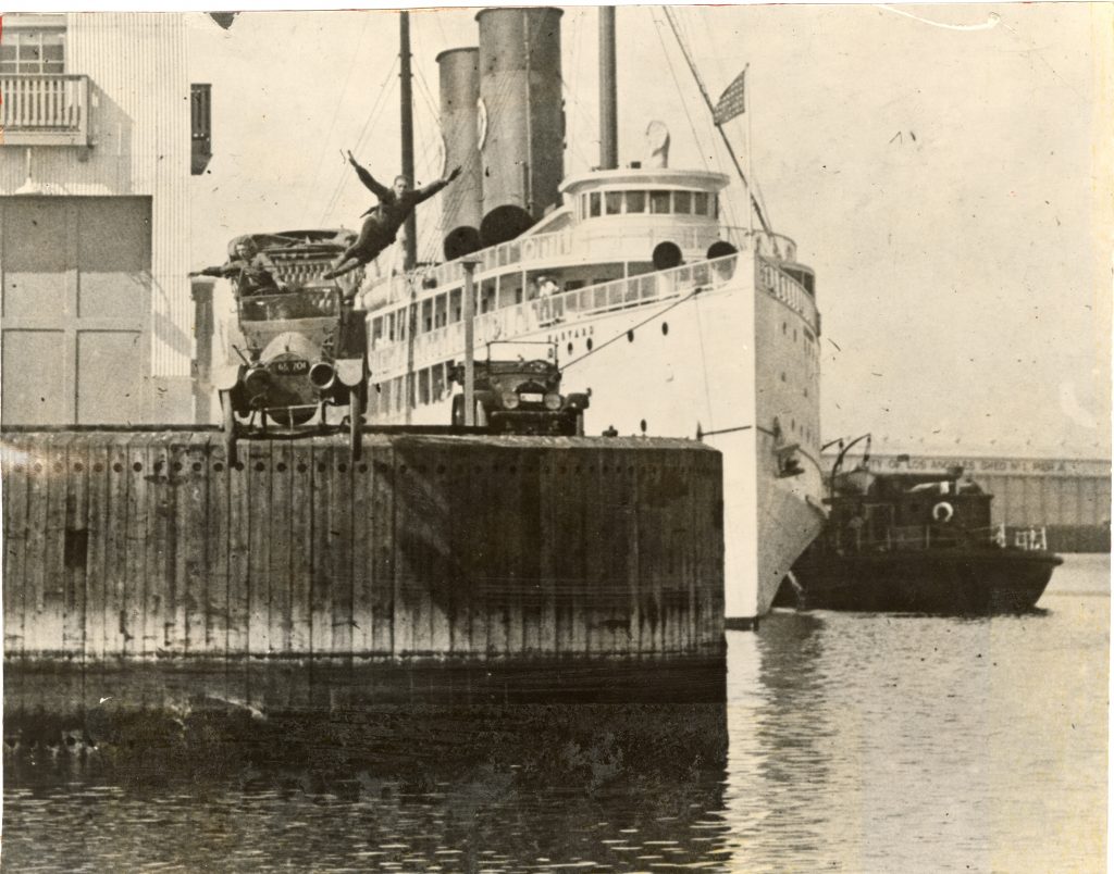 Black and white photograph showing Joe Bonomo jumping out of a car that is falling off a pier toward the water. A woman is in the car. 