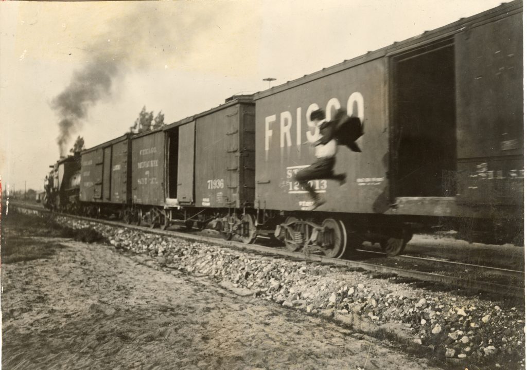 Black and white photograph of Joe Bonomo jumping out of box car of a moving train. Three men on horseback appear to be on top of the train further away.