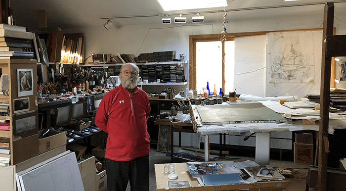 An elderly-aged Igor Galanin wearing a red quarter-zip sweater stands in his art studio in Millwood New York.