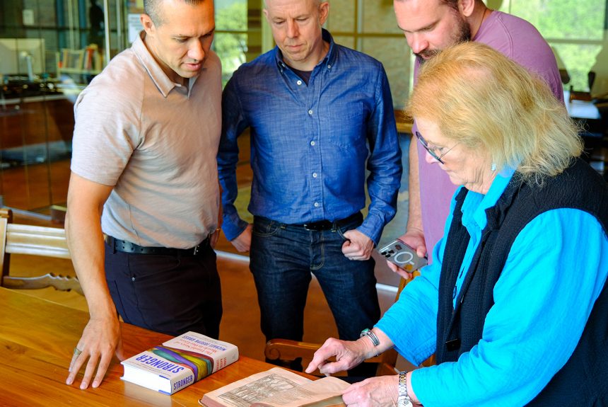 Dr. Charles Stocking, Michael Joseph Gross, Martins Licis, and Dr. Jan Todd look at an artifact on a wood table in the Stark Center reading room.