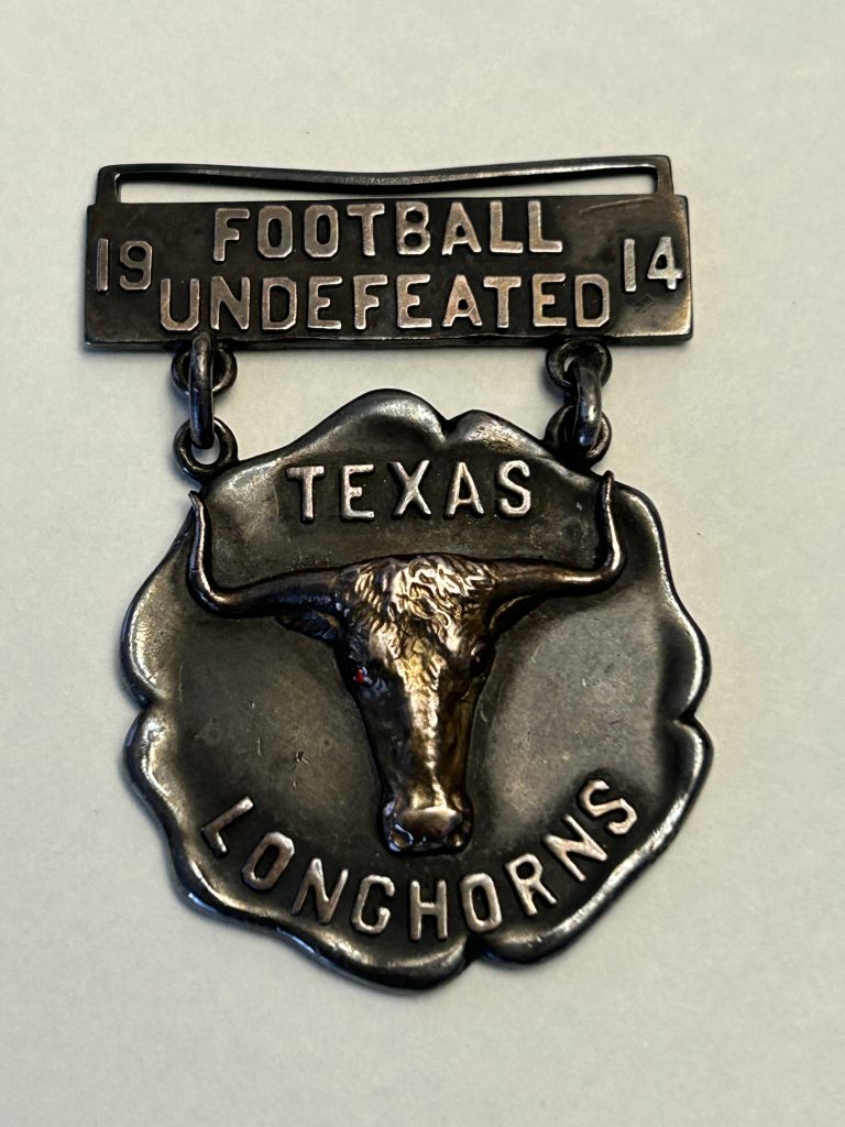 A photo of a silver medal with a longhorn head and the words, "1914 Football Undefeated / Texas Longhorns."